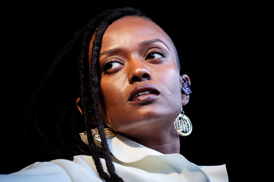 Kelela performing on an outdoor stage in daylight, one hand raised toward the microphone
