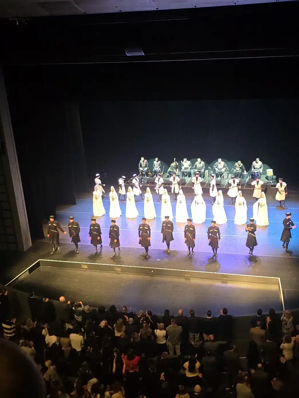 The full company in lines across the stage taking a bow while the audience in the foreground stands and applauds