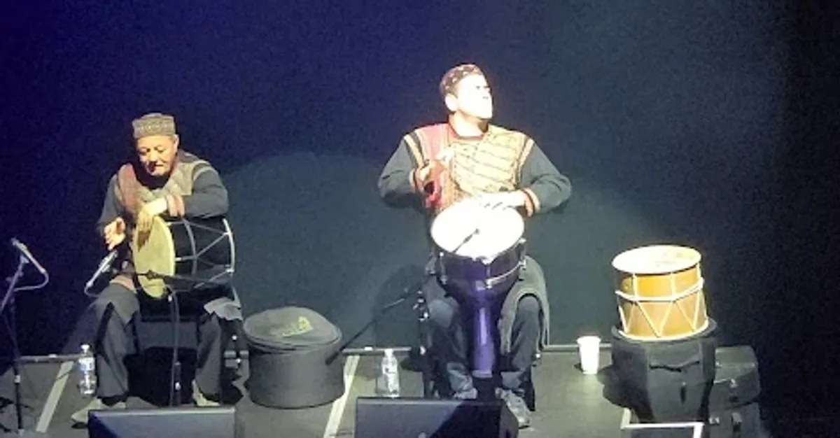 Two drummers seated on a lit platform, both mid-strike on frame drums held across the knee, a third drum standing on a case beside them, the rest of the stage black