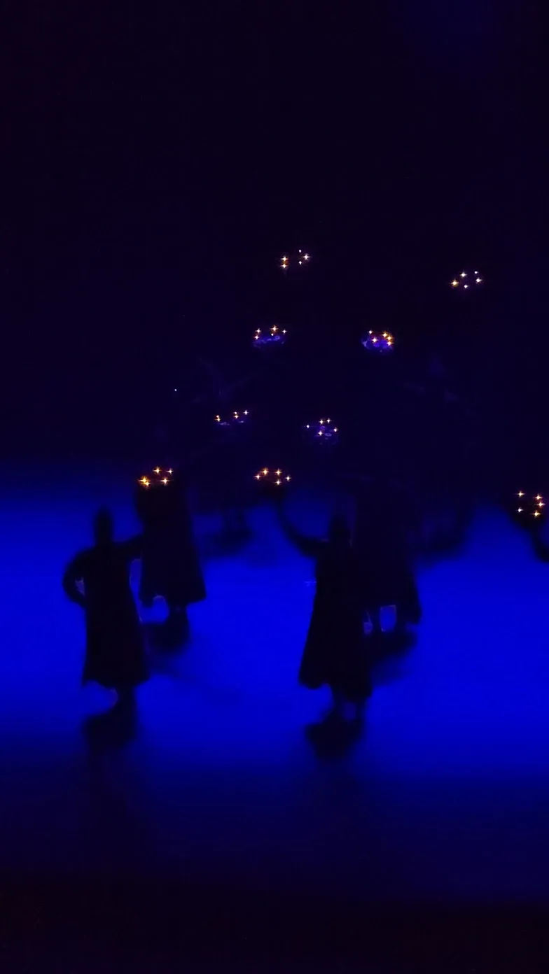 A darkened stage lit only by about a dozen clusters of small warm lights at head height, with dancers silhouetted against deep blue
