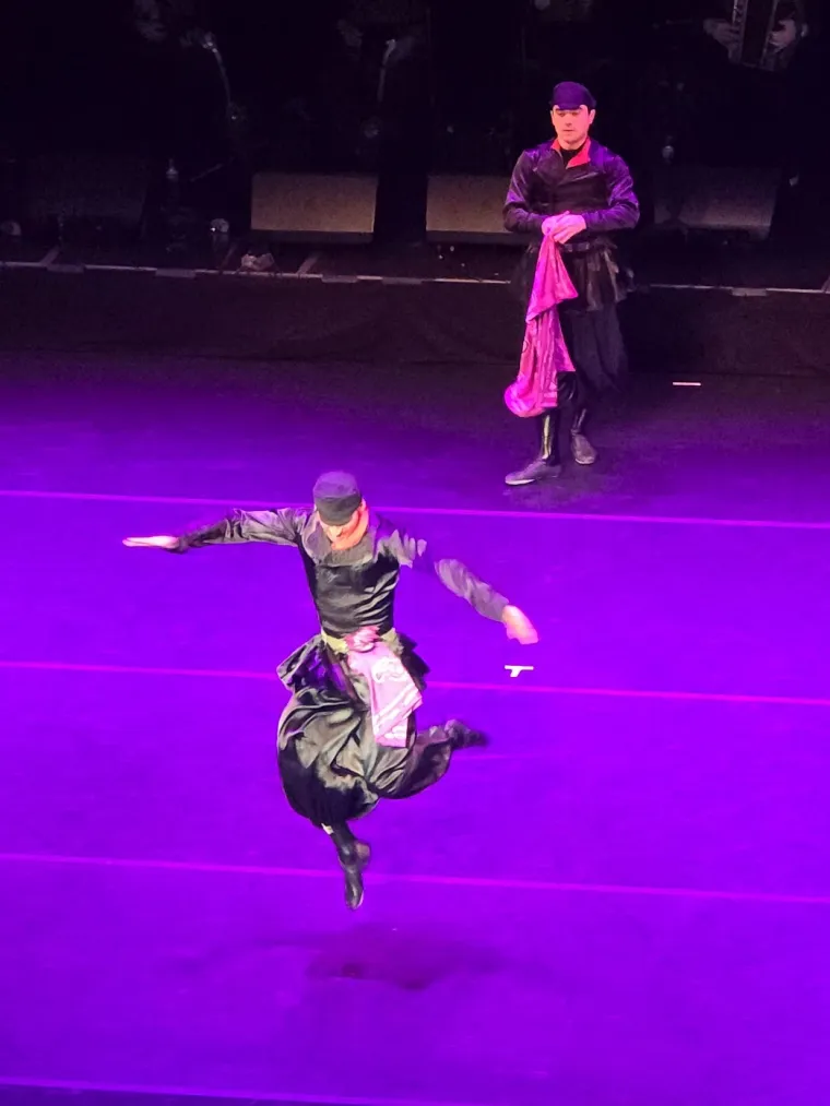 A dancer in a dark baggy costume leaps with arms spread while another stands behind holding a pink silk shawl, under purple light