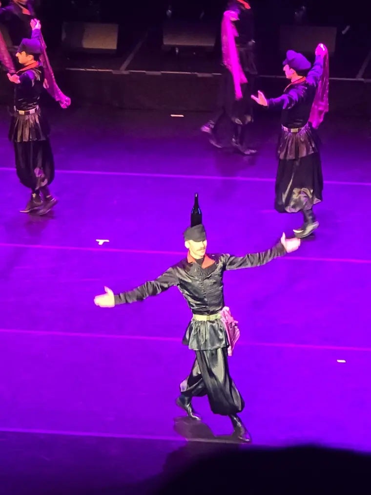 A dancer walks forward with arms open and a wine bottle balanced upright on his cap while others move behind him in purple light