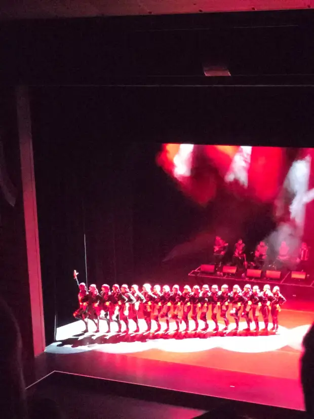 A long line of dancers in red and dark costume bent in unison under red light, musicians on a platform behind