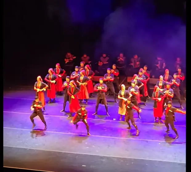 A mixed ensemble in red and black costume mid-step under purple and blue light with musicians behind