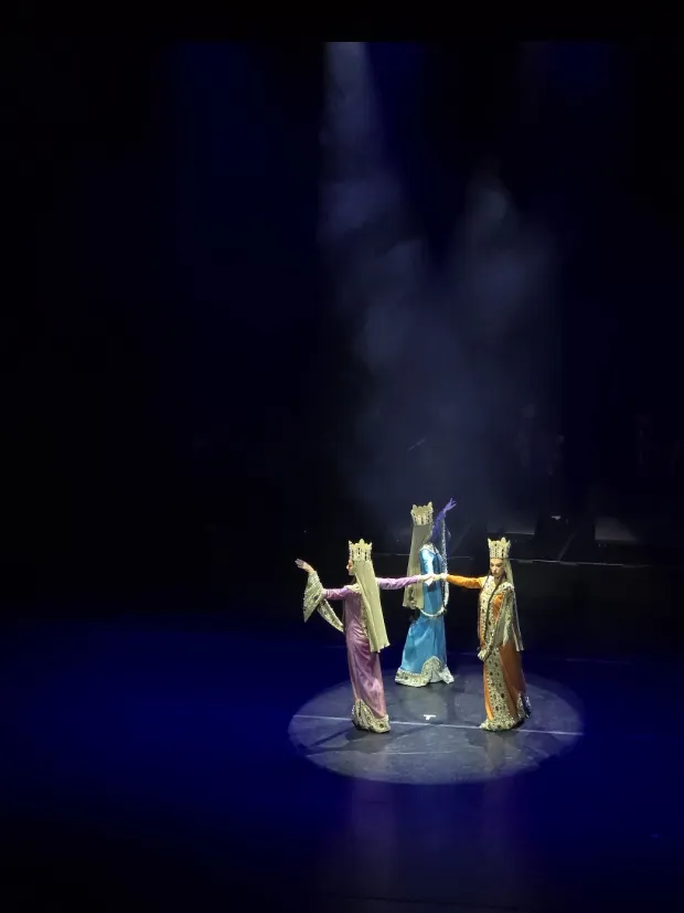 Three dancers in long gowns and tall gold crowns stand close together in a spotlight, arms extended