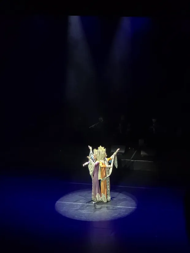 The same three crowned dancers drawn into a tight column in a spotlight, arms raised