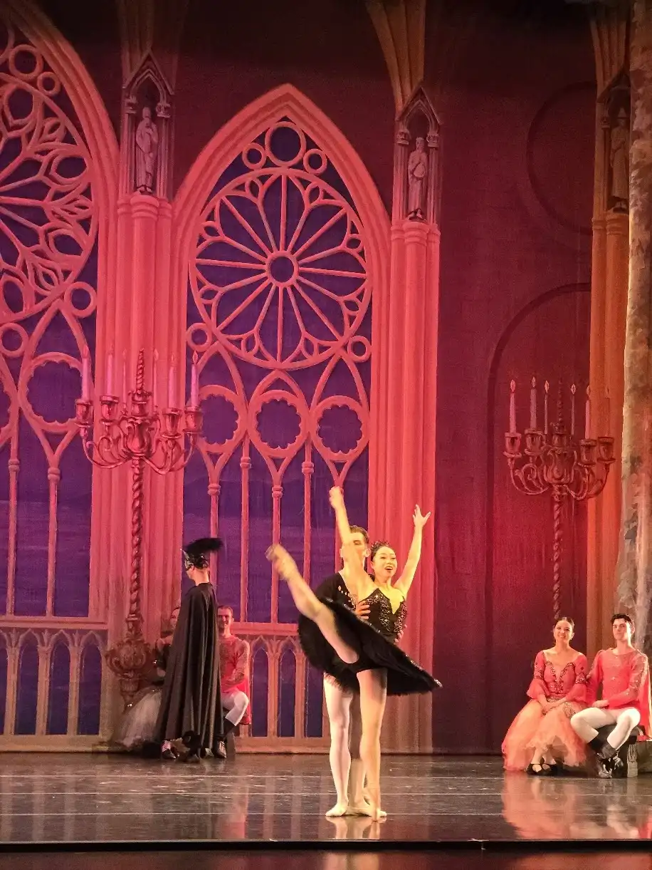 A ballerina in a black tutu is supported in arabesque before a coral-lit gothic rose window, the court seated around her