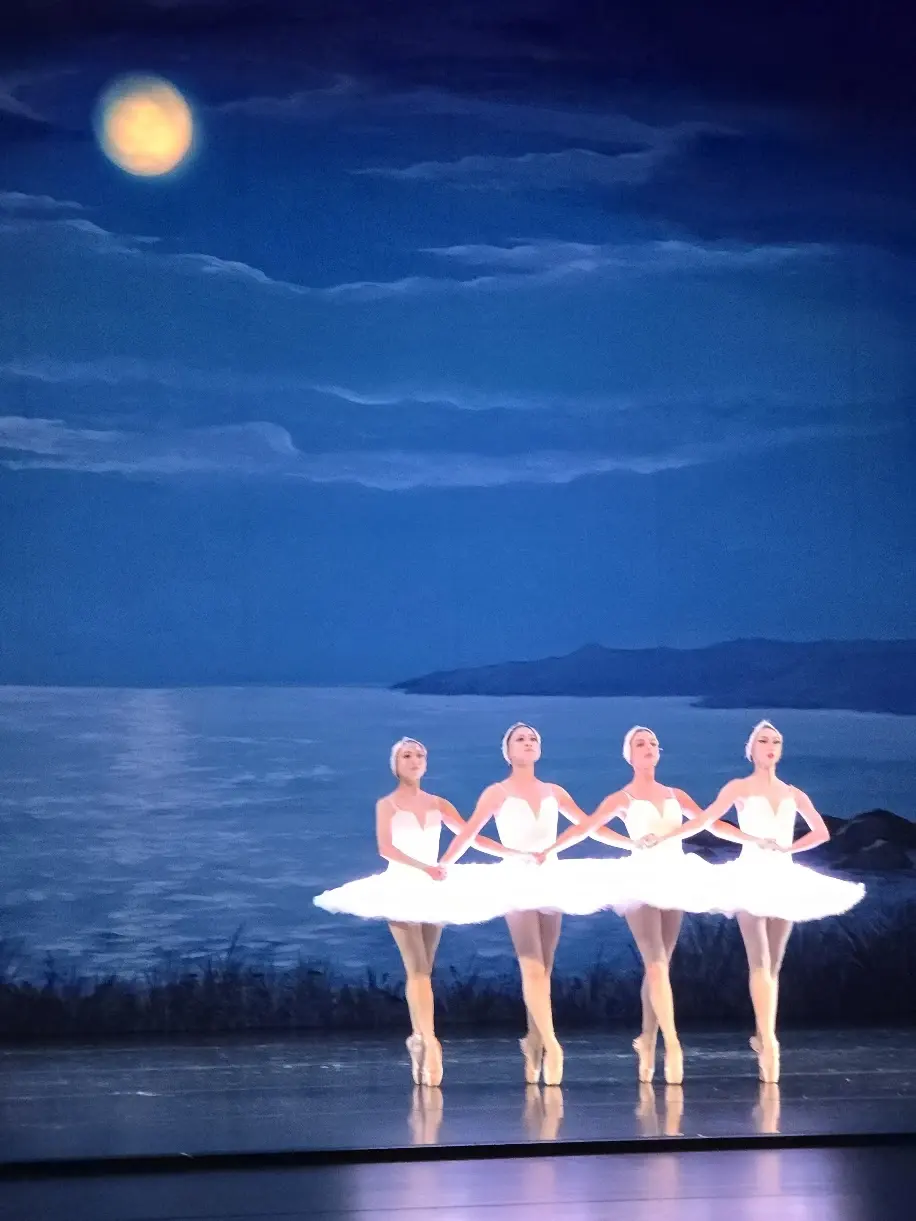 Four dancers in plain white tutus linked hand to hand in a line, dancing the pas de quatre
