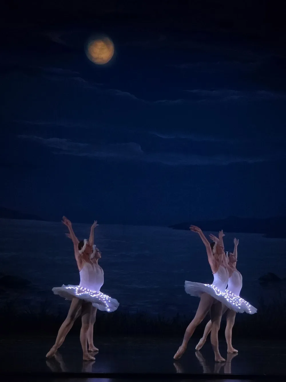 Four dancers in illuminated tutus with arms raised beneath a painted full moon on a darkened stage
