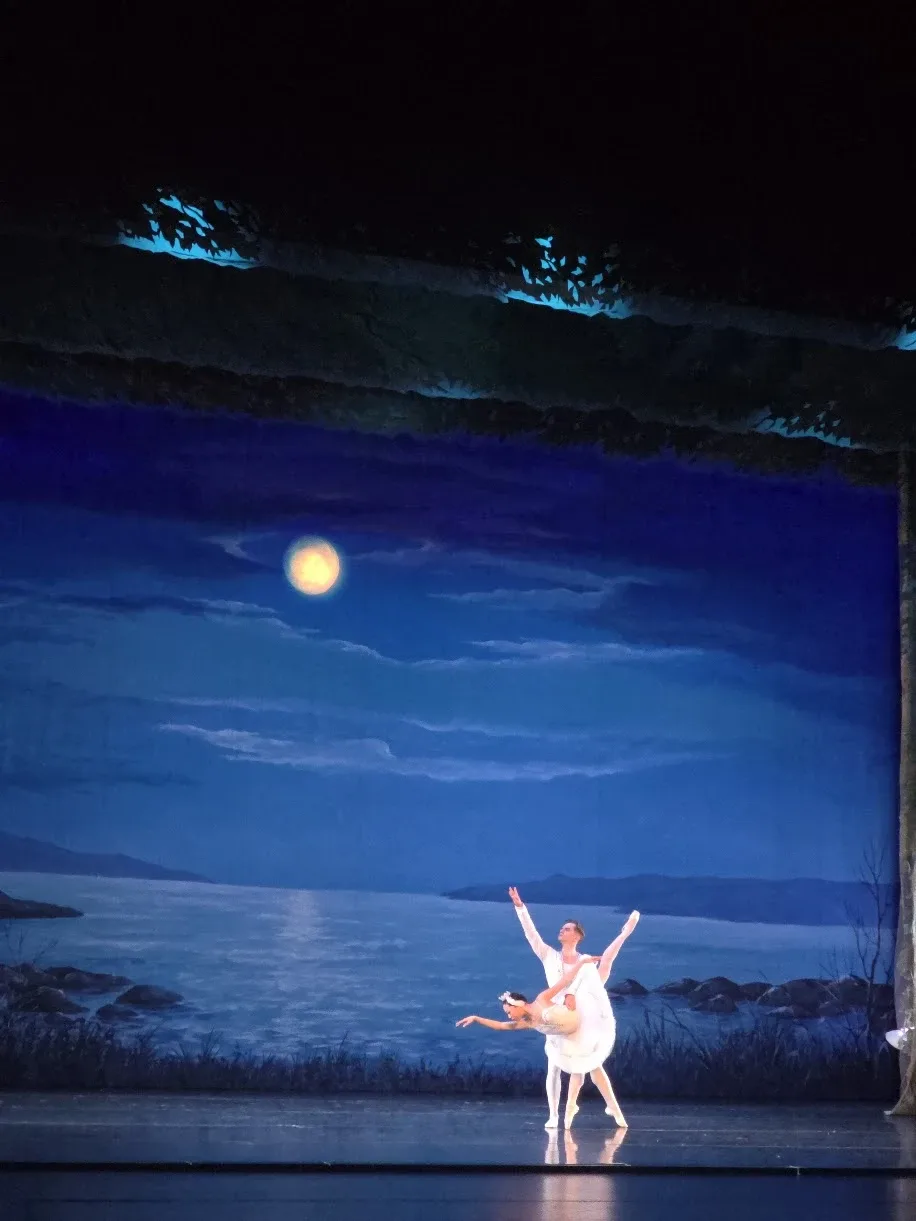 A dancer in white supports a ballerina in a deep backbend beneath a painted moon on a dark stage