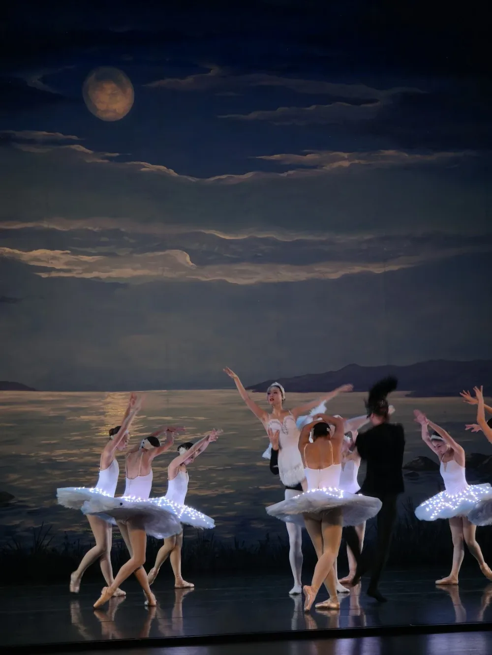 A figure in black with a feathered headdress crosses in front of dancers in illuminated tutus while a swan is lifted behind him